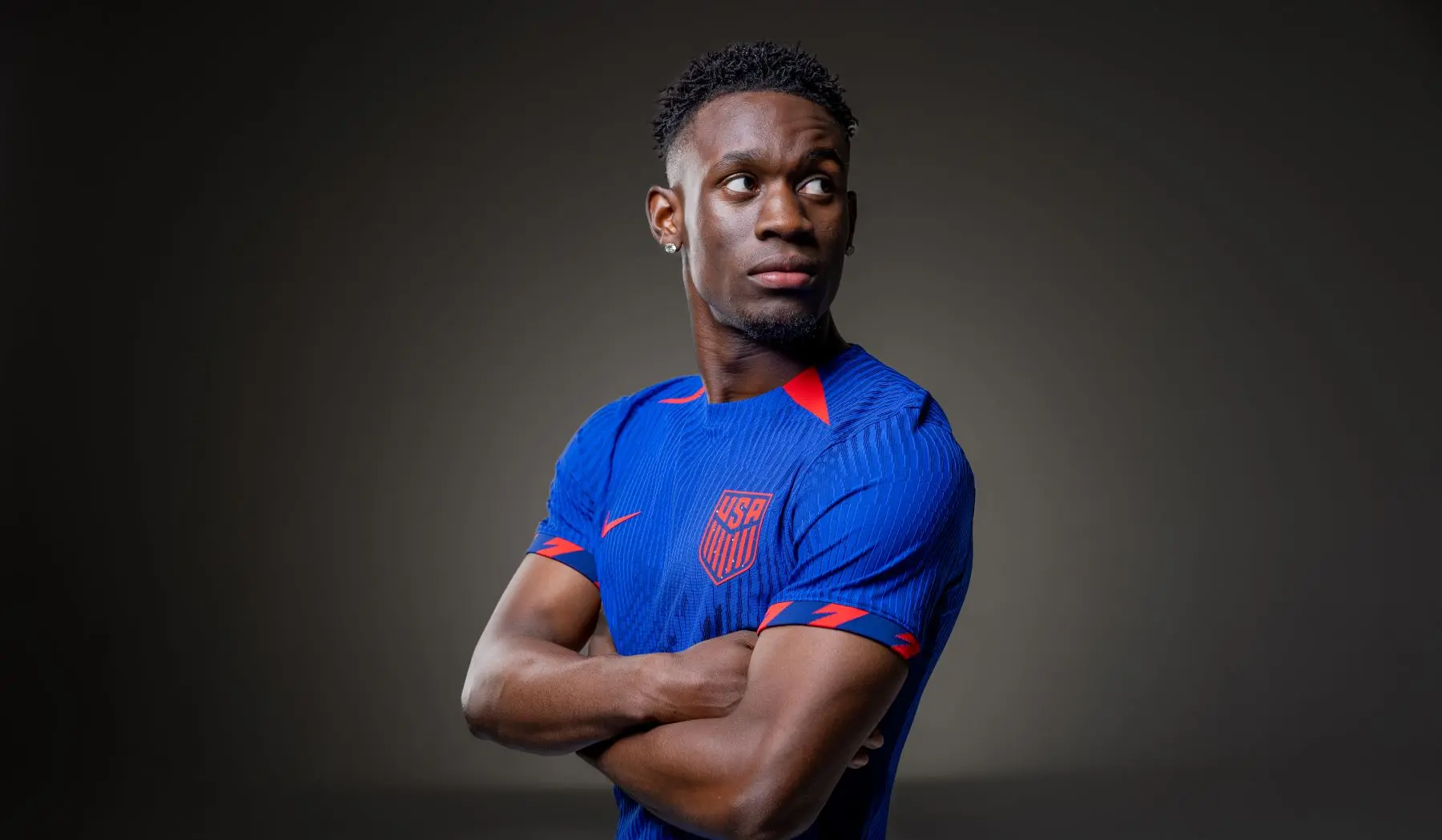 Folarin Balogun in a blue USA soccer jersey, arms crossed, looking to the side.