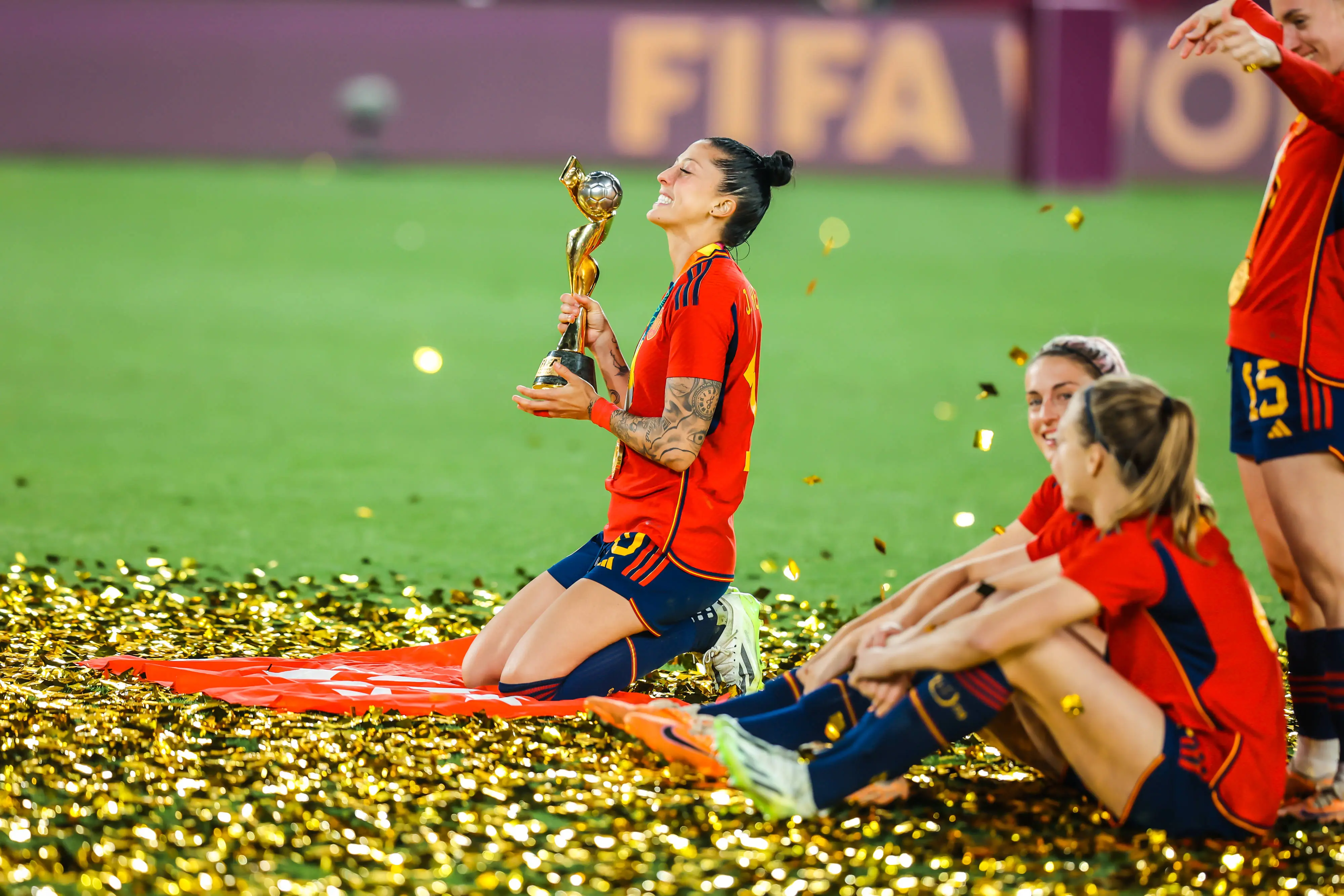 A female football player from Spain celebrating with a trophy surrounded by teammates.